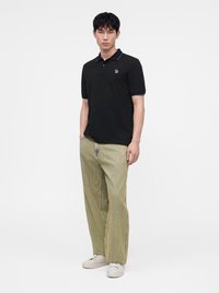 Homme portant un polo noir avec une bordure bleue, un pantalon large vert clair et des baskets blanches, debout avec une main dans la poche.