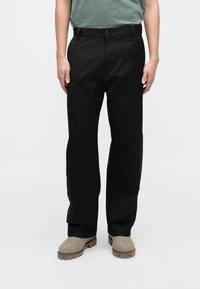 Schwarze, weit geschnittene Hose aus strapazierfähigem Material, mit seitlichen Taschen und flacher Front. Trägt beige Schuhe.