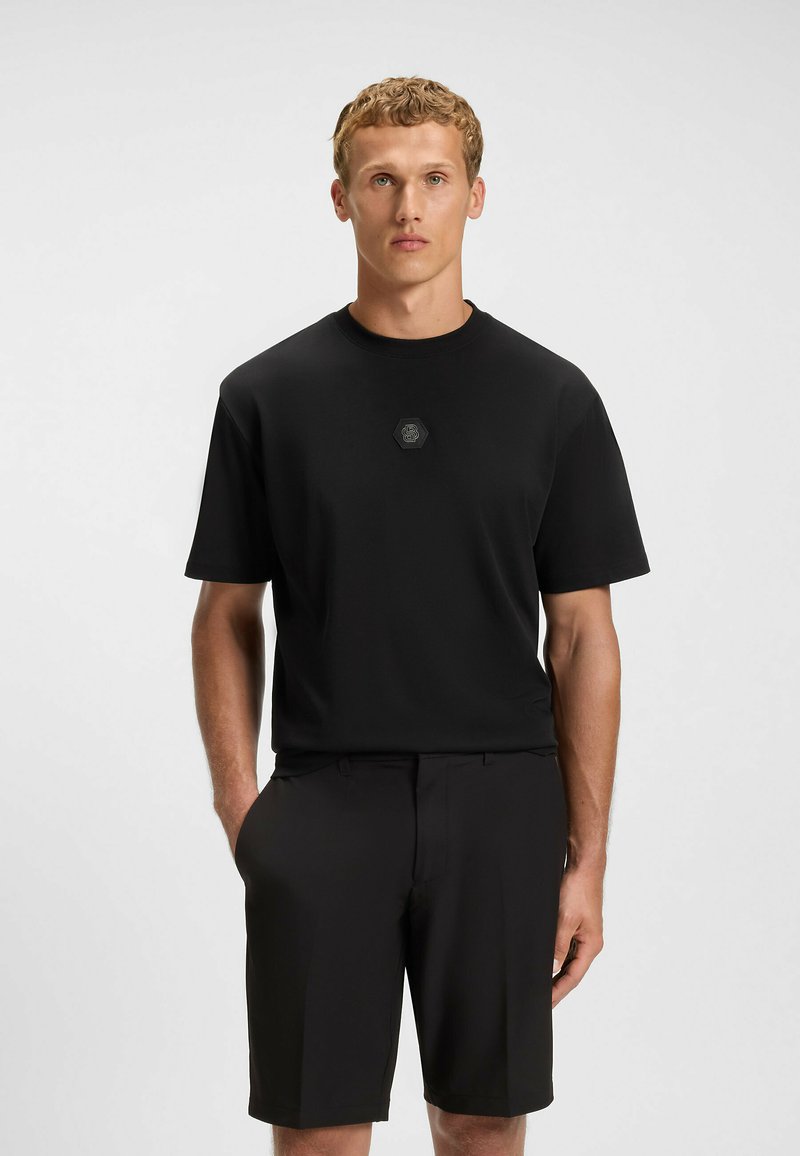 Czarna koszulka z krótkim rękawem, okrągłym dekoltem i sześciokątną łatą z logo na piersi, w połączeniu z czarnymi szortami. Gładka faktura materiału.