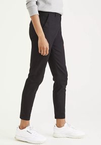 Čierne slim-fit nohavice s hladkou textúrou, s stredne vysokým pásom a postrannými vreckami, v kombinácii s bielymi športovými teniskami.