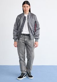 Grijze bomberjack met geribbelde manchetten, wit T-shirt, verweerde grijze spijkerbroek en zwarte sneakers met witte zolen en blauwe accenten.