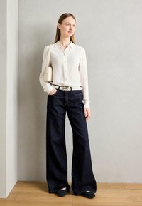 Fehér, hosszú ujjú, gombos ing, sötétkék, magas derekú, bő szárú farmernadrág, fekete slip-on cipő, és egy kis bézs kézitáska.