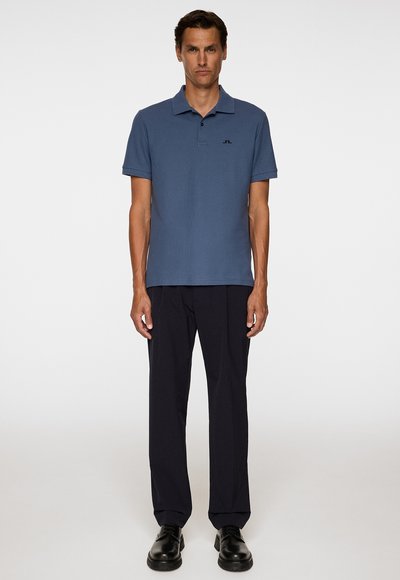 Blauwe polo met een kraag en korte mouwen, voorzien van een klein zwart logo, gecombineerd met donkere broek en zwarte schoenen.