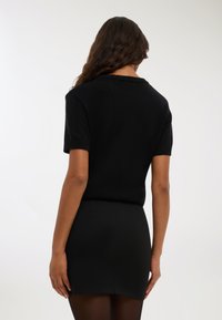 Kobieta z długimi falującymi włosami, ubrana w czarną, krótką bluzkę dzianinową i czarną mini spódniczkę, stojąca na jednolitym jasnym tle.