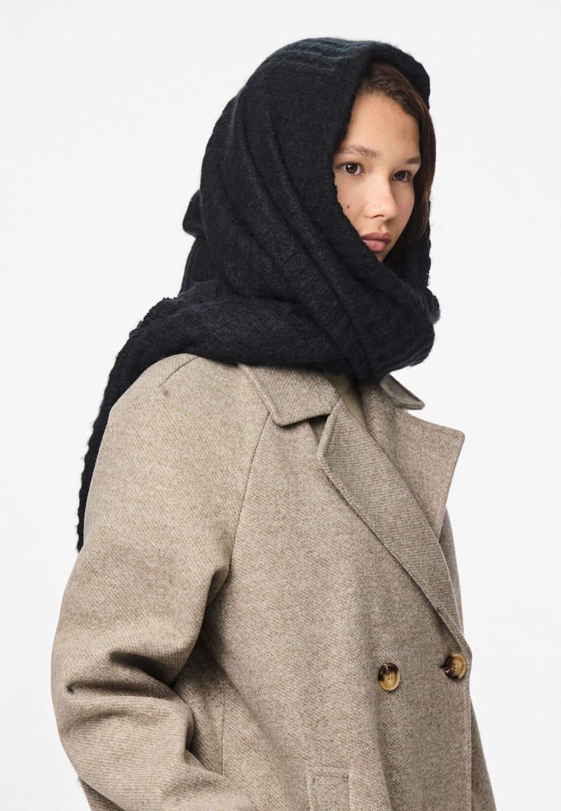 Sort, struktureret uldscarf draperet over hovedet, parret med en lys beige, dobbeltbrystet frakke med guldknapper og et blødt stof.