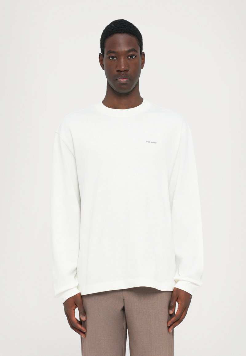 White sweatshirt with long sleeves, round neck, and embroidered logo. Lightweight material with a smooth texture. Paired with beige trousers.