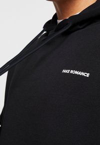 Felpa nera in cotone con finitura testurizzata, caratterizzata da un logo ricamato bianco "FAKE ROMANCE" sul petto e coulisse regolabili.