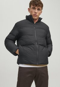 Schwarze Pufferjacke mit hohem Kragen, Reißverschluss und breitem Steppdesign. Model steht mit den Händen in den Taschen und trägt ein geschichtetes Outfit.