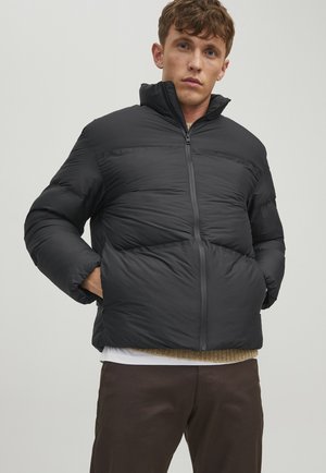 Sort pufjakke med høj krave, lynlås og bredt quiltet design. Modellen står med hænderne i lommerne iført et lagdelt outfit.