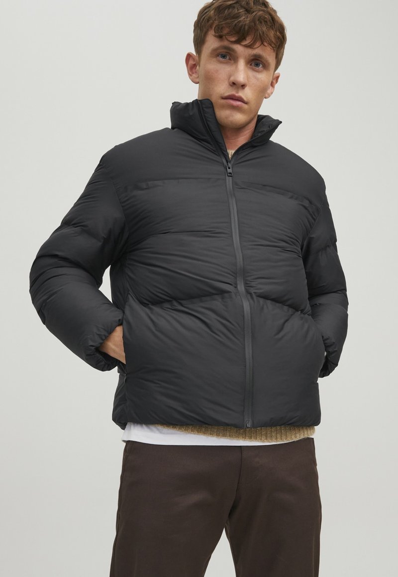 Schwarze Pufferjacke mit hohem Kragen, Reißverschluss und breitem Steppdesign. Model steht mit den Händen in den Taschen und trägt ein geschichtetes Outfit.