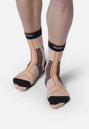 Gambe che indossano calzini sportivi beige con motivi geometrici neri, arancioni e bianchi e punte nere su sfondo bianco uniforme.