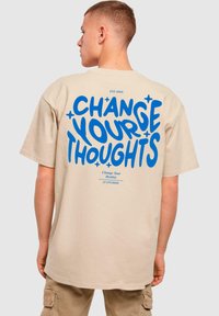 Beige bomulds t-shirt med løs pasform, med fed blå tekst "ÆNDRE DINE TANKER" og accenter, trykt på ryggen.