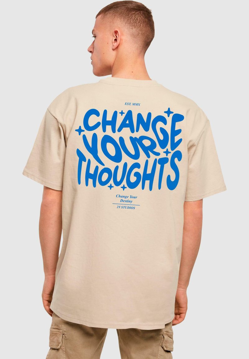 Beige bomulds t-shirt med løs pasform, med fed blå tekst "ÆNDRE DINE TANKER" og accenter, trykt på ryggen.