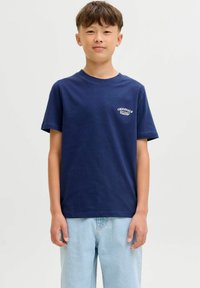 Navyblauwe katoenen t-shirt met korte mouwen, ronde hals, met een witte logo graphic op de linkerkant van de borst. Draagt het met lichtblauwe jeans.