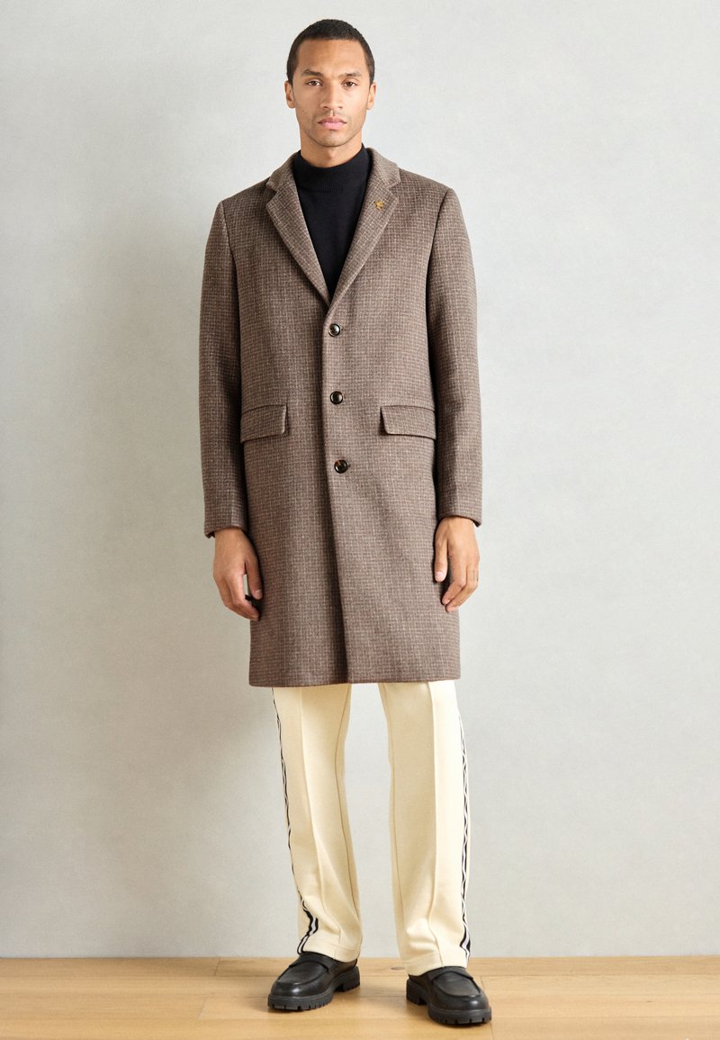 Abrigo de lana a cuadros marrones con solapa, botones frontales y bolsillos laterales, combinado con pantalones blancos con rayas negras y zapatos negros gruesos.