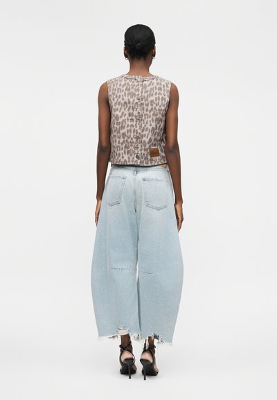 Γυναίκα με σκούρο δέρμα και μαλλιά σε κότσο που φοράει αμάνικη μπλούζα με λεοπάρ μοτίβο και κουμπιά στην πλάτη, φαρδιά ανοιχτό μπλε τζιν και μαύρα τακούνια.