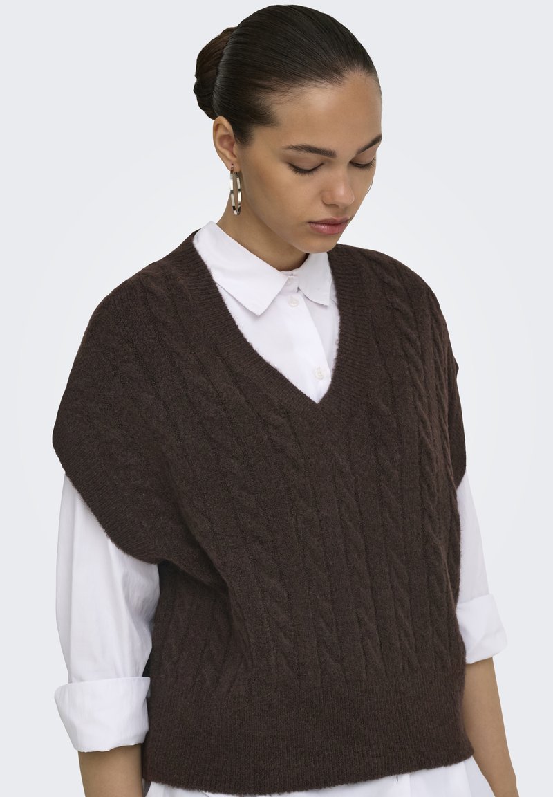 Femme portant un pull sans manches en maille torsadée marron foncé par-dessus une chemise blanche à col avec manches retroussées, regardant vers le bas.
