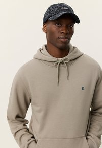 Ανοιχτό μπεζ φούτερ με κορδόνι και μικρό μπλε λογότυπο στο στήθος. Φορεμένο με σκούφο ναυτικού χρώματος με λουλουδάτο σχέδιο. Λεία υφή.