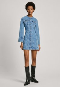 Denim kleita ar garām piedurknēm, pogu priekšpusi un divām krūšu kabatām. Gaiši zila krāsa ar pieguļošu dizainu un izplestu svārku. Melni ceļgalu augstie zābaki.