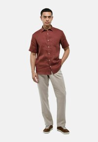 Chemise en lin bordeaux à manches courtes avec une poche poitrine, devant boutonné, associée à un pantalon beige clair et des baskets marron.