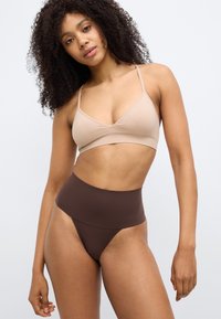 Μπεζ ασύρματο σουτιέν με λεπτές τιράντες, συνδυασμένο με καφέ ψηλόμεσο στρινγκ. Λείος υφασμάτινος ιστός, μινιμαλιστικός σχεδιασμός, χωρίς ορατά μεταλλικά στοιχεία.