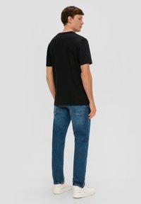 T-shirt en coton noir à manches courtes, col rond, associé à un jean en denim bleu et des baskets blanches, vu de dos.