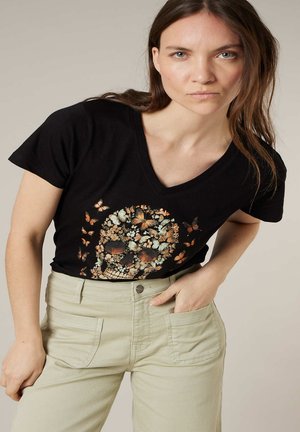 Femme portant un t-shirt noir à col en V avec un motif de crâne fleuri et de papillons, assorti à un pantalon beige clair taille haute, main dans la poche.