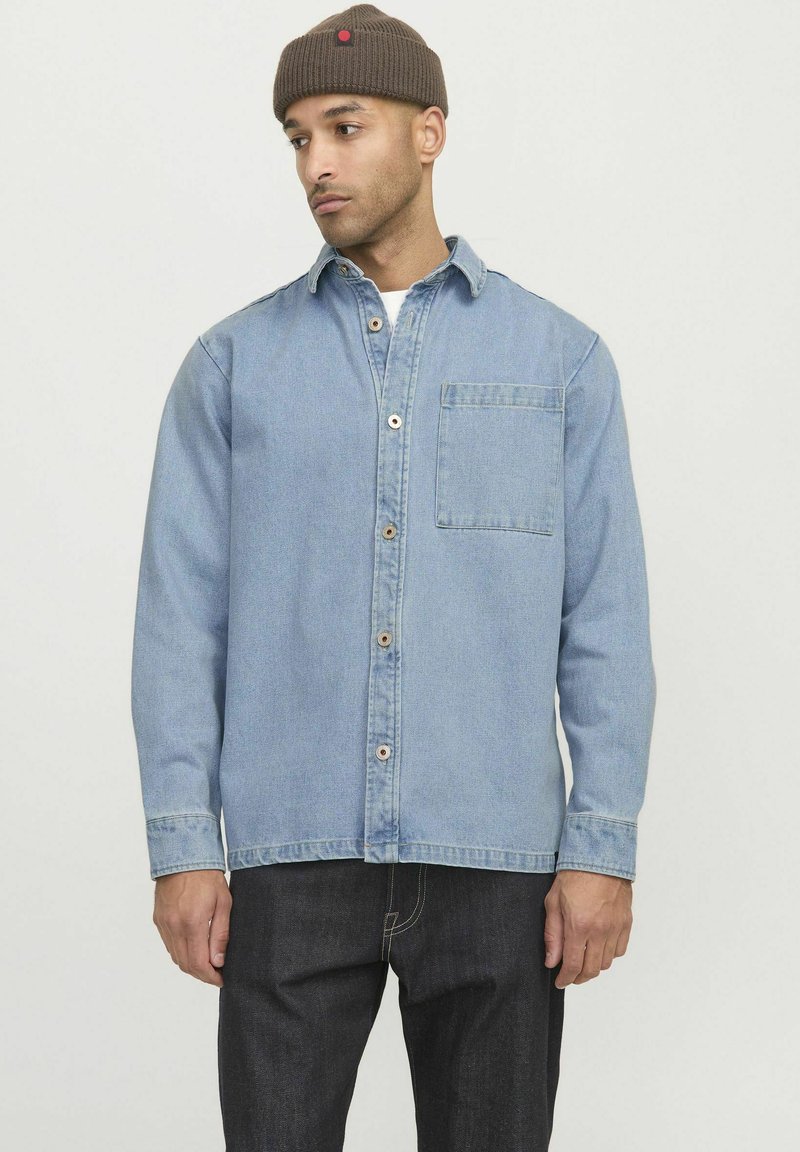 Camicia in denim blu chiaro con chiusura a bottoni, colletto e taschino sul petto. Maniche lunghe con polsini, abbinata a pantaloni in denim scuro.
