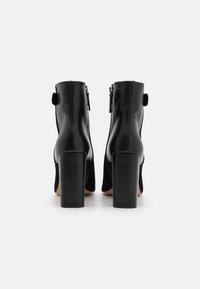 Czarne skórzane botki za kostkę na wysokim obcasie w kształcie słupka, z zamkiem błyskawicznym oraz dekoracyjną zakładką z tyłu. Gładka tekstura, elegancki design.