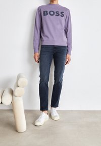 Felpa lilla con la scritta "BOSS" in grassetto, abbinata a jeans di denim scuro e sneakers bianche con accenti chiari. Tessuto liscio, vestibilità casual.