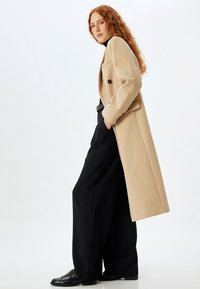 Lungo cappotto beige su un dolcevita nero e pantaloni neri a gamba larga, con un solo bottone, tasche e mocassini neri con accenti in metallo.