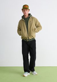 Fleece jacket in olive green, zippered front, two pockets; paired with a green hoodie, black pants, and gray sneakers with green accents.