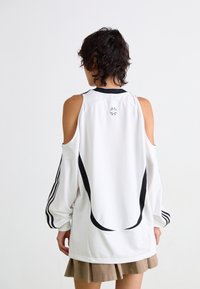 Witte pullover met cold-shoulder ontwerp, zwarte zijaccenten en een klein logo op de achterkant. Lange, volumineuze mouwen. Lichtgewicht materiaal.
