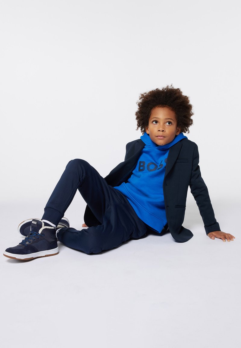 Marineblazer over een blauwe hoodie met "BOSS" opdruk, gecombineerd met marine joggingsbroek en blauwe sportschoenen. Gelaagd, casual ontwerp.