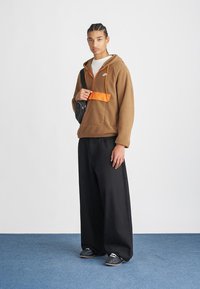 Mladý muž s vrkôčkami vo vlasoch, oblečený v hnedom mikine, širokých čiernych nohaviciach, čiernych topánkach a nesúci čierny crossbody batoh na modrej podlahe.