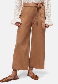 Pantalons à jambes larges en daim marron avec une ceinture à nouer, dotés de poches latérales et d'une texture lisse. Associés à des chaussures plates à brides marron.