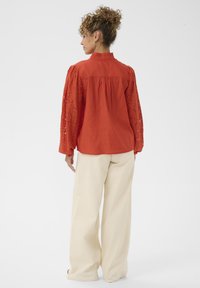 Blouse rouge à manches longues en dentelle, col mandarin, et détail froncé au dos, portée avec un pantalon ample crème.