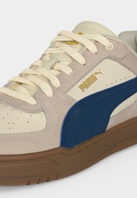 Μπεζ και κρεμ αθλητικό παπούτσι Puma με ναυτικό μπλε πλαϊνή ρίγα και καφέ σόλα, με κρεμ κορδόνια και χρυσό λογότυπο Puma στο πλάι.