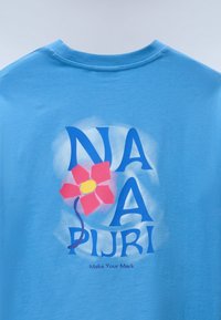 T-shirt bleu avec de grandes lettres bleues affichant "NA A PIJRI" autour d'une fleur rouge à centre jaune et la phrase "Make Your Mark" en dessous.