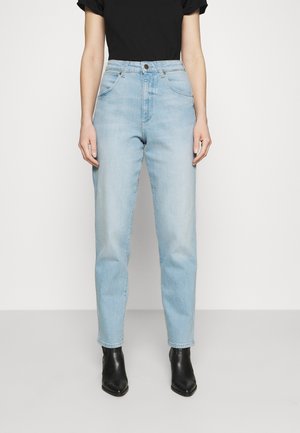 Osoba ubrana w jasnoniebieskie dżinsy z wysokim stanem, czarny krótki top i czarne botki, stoi na jednolitym tle.