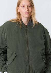 Jeune femme aux cheveux blonds portant une veste bomber zippée oversize verte sur fond blanc uni.