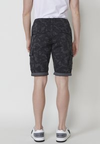 Shorts cargo noirs à motif camouflage, avec une taille élastique, un ourlet retroussé, plusieurs poches latérales et un tissu texturé. Portés avec des baskets blanches.