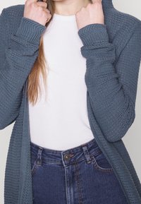 Lange openvallende cardigan in een gestructureerde leigrijze breisel. Heeft geribde manchetten en een relaxte pasvorm, gecombineerd met een getailleerd wit shirt en denim.