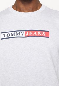 Šedá mikina s kulatým výstřihem; má horizontální pruh s logem v červené, bílé a námořnické barvě s textem "TOMMY JEANS". Měkká textura.