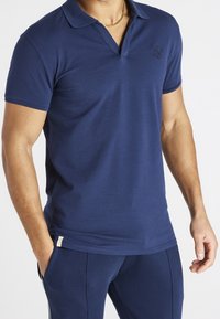 Mies, der ein kurzärmliges Poloshirt in Marineblau mit einem kleinen gestickten Logo auf der Brust und dazu passenden Marineblauen Hosen trägt.