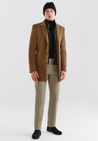 Brązowy płaszcz wełniany z klapami, czarny golf, jasne khaki spodnie, czarne botki za kostkę i fakturowana czarna czapka z dzianiny.