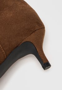 Acercamiento de un tacón de bota de tobillo de ante marrón con tacón curvado negro y punta de goma sobre fondo blanco.