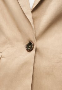 Κοντινή λήψη μπεζ σακακιού κουμπωμένο με ένα μεγάλο καφέ κουμπί, που δείχνει το πέτο και μέρος της τσέπης.