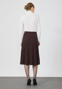 Jupe plissée brune avec une large taille, associée à un chemisier blanc à manches longues. Des escarpins noirs complètent la tenue.
