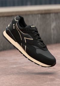 Black athletic shoe with a smooth leather upper, perforated accents, white midsole, and distinctive logo detail. Textured laces and sole.